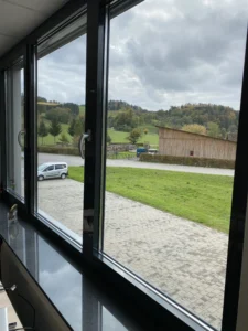 Blick aus einem Büro mit großen Fenstern auf eine grüne Landschaft mit Hügeln und Bauernhof.