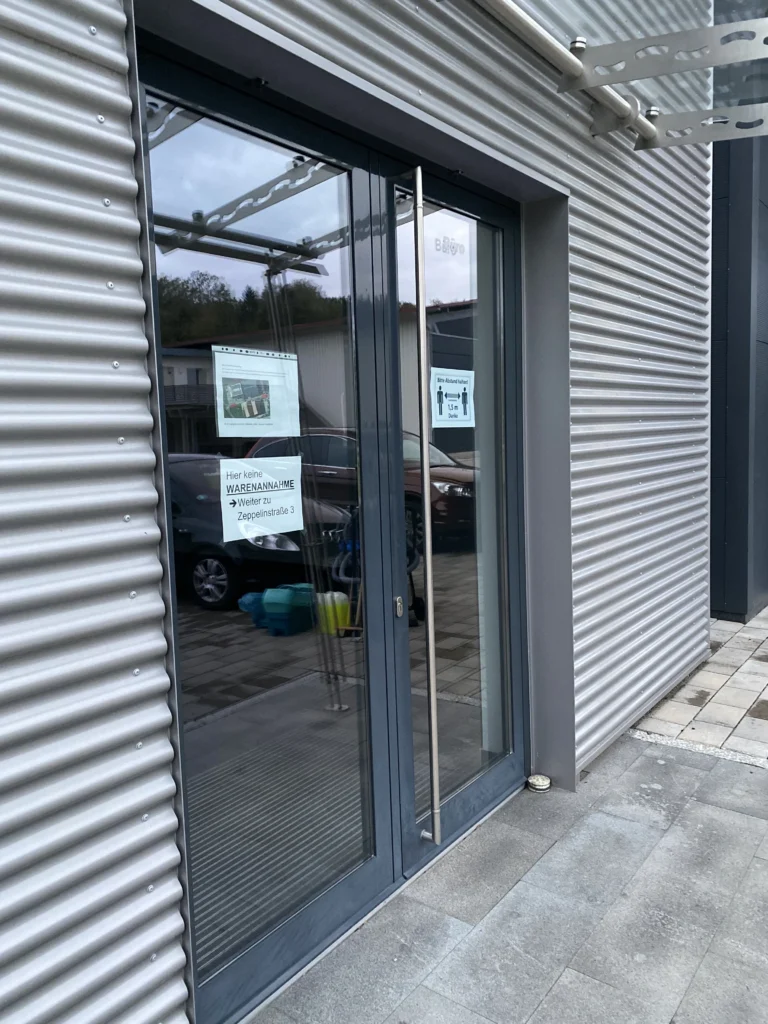 Büroeingang mit modernen Glastüren, Hinweisschildern und metallischer Fassade.