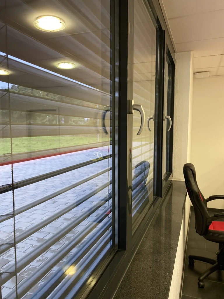 Bürofenster mit geschlossenen Außenjalousien, schwarzen Fensterrahmen und Granitfensterbank.