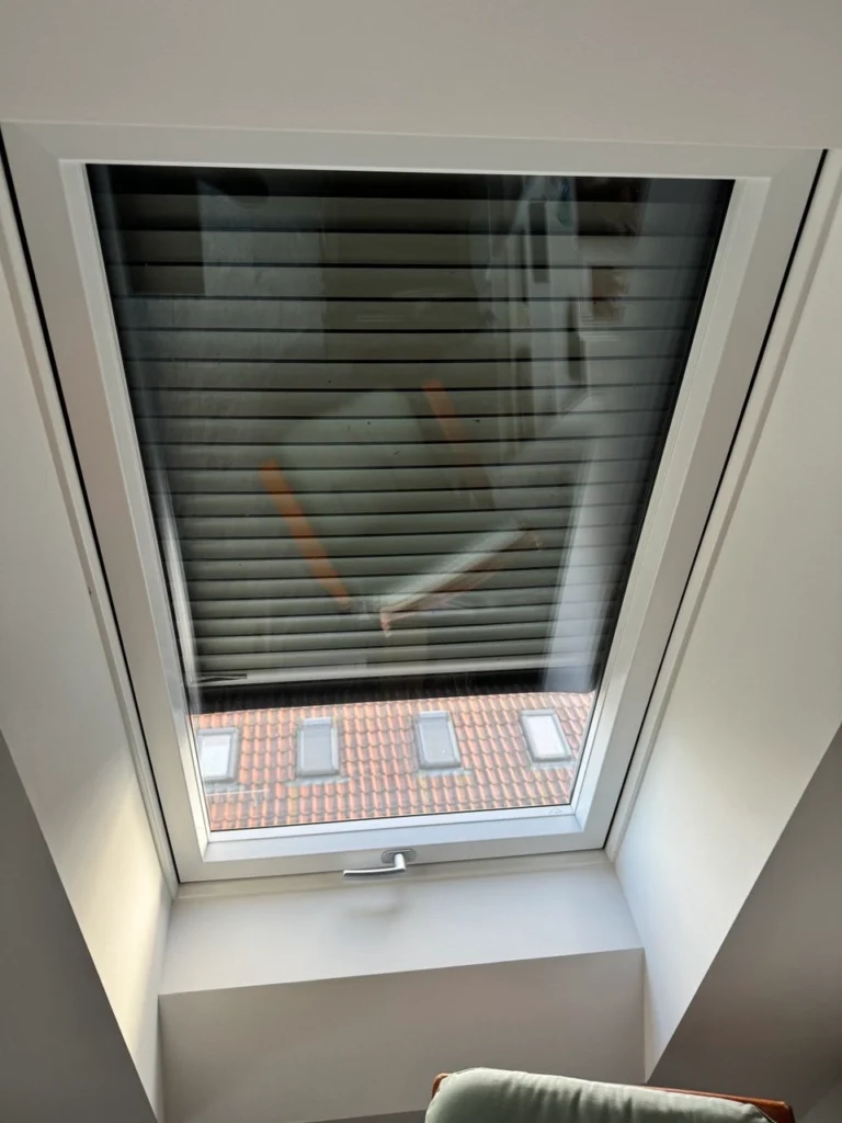Dachfenster mit geschlossenem Außenrollo, das die Reflexion eines Stuhls und eines roten Ziegeldachs zeigt.