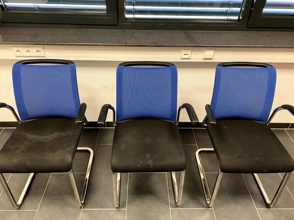 Drei Bürostühle mit blauer Rückenlehne und schwarzem Sitz, aufgestellt vor einem Fenster.