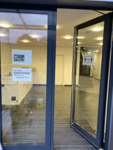 Eingang eines Bürogebäudes mit geöffneter Glastür, Hinweisschildern und Blick auf den Empfangsbereich.