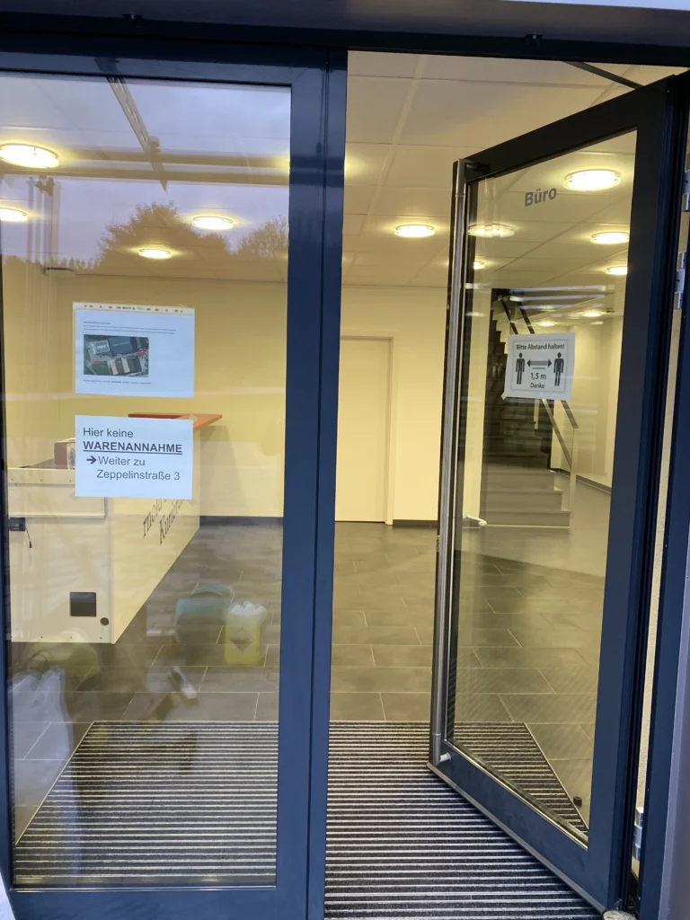 Eingang eines Bürogebäudes mit geöffneter Glastür, Hinweisschildern und Blick auf den Empfangsbereich.