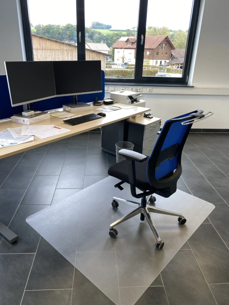 Moderner Büroarbeitsplatz mit höhenverstellbarem Schreibtisch, zwei Monitoren, ergonomischem Drehstuhl und einer Bodenschutzmatte.