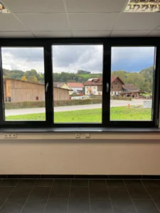 Großes Bürofenster mit Blick auf eine ländliche Gegend mit Holzhäusern und grünen Wiesen.