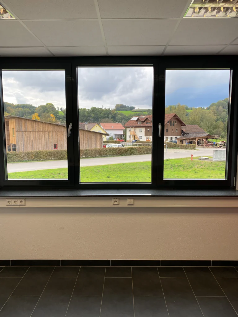 Großes Bürofenster mit schöner Aussicht Großes Bürofenster mit Blick auf eine ländliche Gegend mit Holzhäusern und grünen Wiesen.