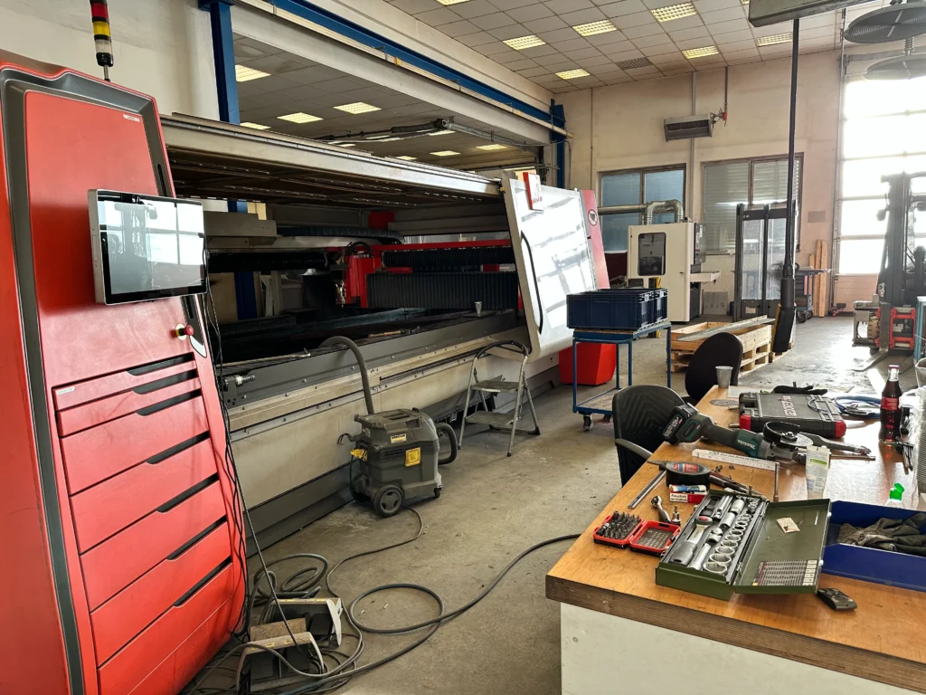 Laserschneidanlage für die Metallbearbeitung in einer Industriehalle mit Werkbänken, Werkzeugen und moderner Maschinentechnik.