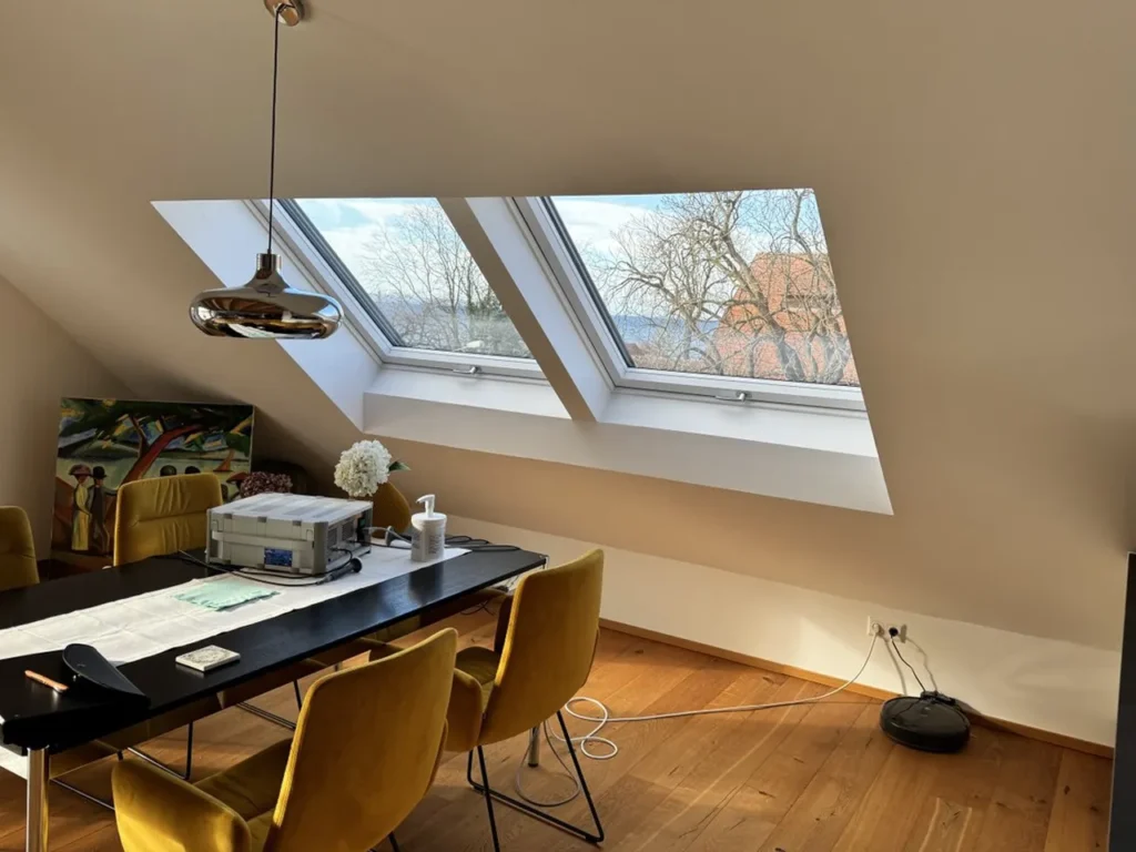 A modern attic office with large skylights, a sleek black dining table, mustard yellow chairs, and a contemporary pendant light, filled with natural light.