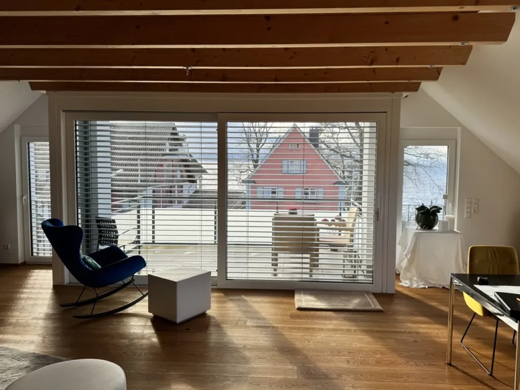 A modern living room with wooden ceiling beams, large windows with blinds, and a cozy interior overlooking a snow-covered suburban house.