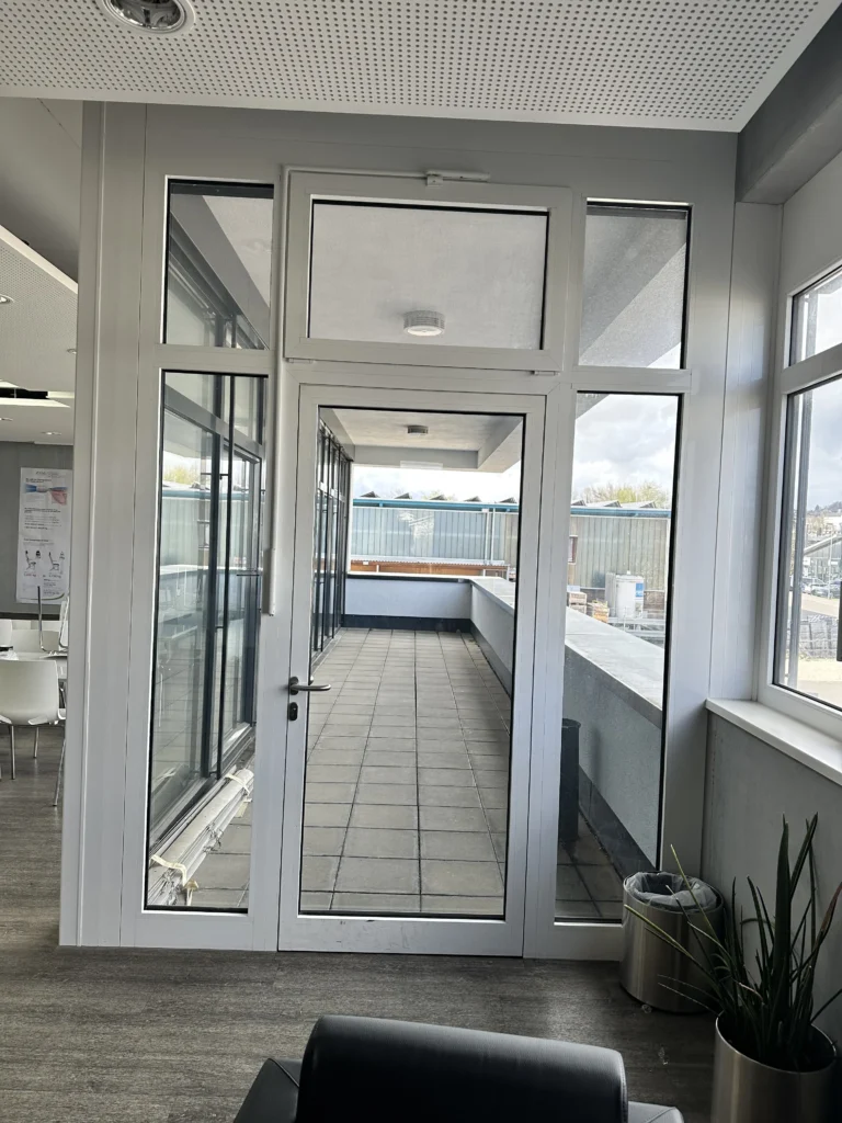 Moderne Glastür mit Zugang zur Terrasse eines Gewerbegebäudes, umgeben von großen Fenstern und einer hellen Innenraumgestaltung.