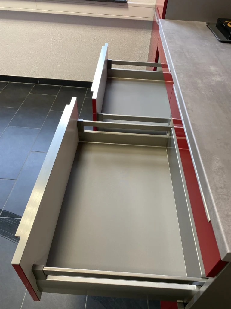 Zwei geöffnete moderne Küchenschubladen mit grauer Innenfläche und roten Fronten in einer Büro-Küche.