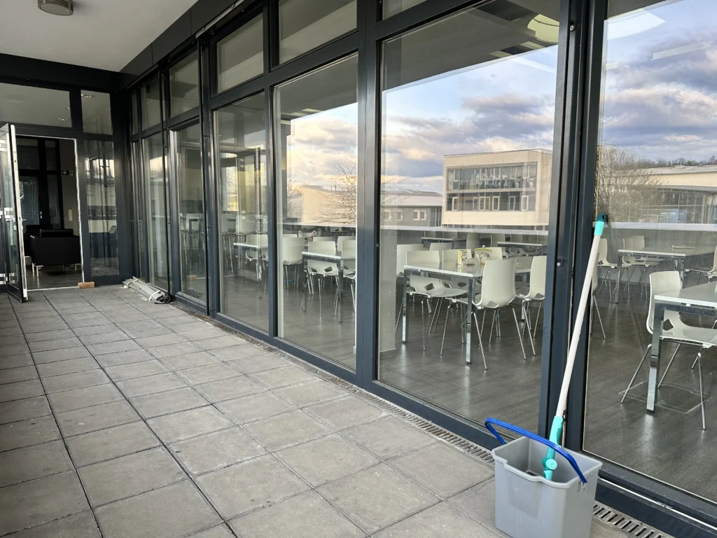 Moderne Terrasse eines Gewerbegebäudes mit großen Glasfronten, die den Blick in einen Speisesaal mit weißen Stühlen und Tischen freigeben.