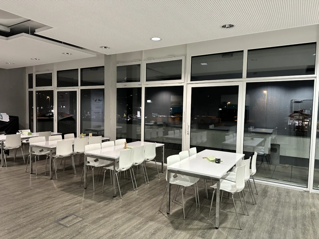 Moderner Speisesaal mit großen Glasfenstern und Blick auf die beleuchtete Stadt bei Nacht. Weiße Tische und Stühle sorgen für eine minimalistische Atmosphäre.