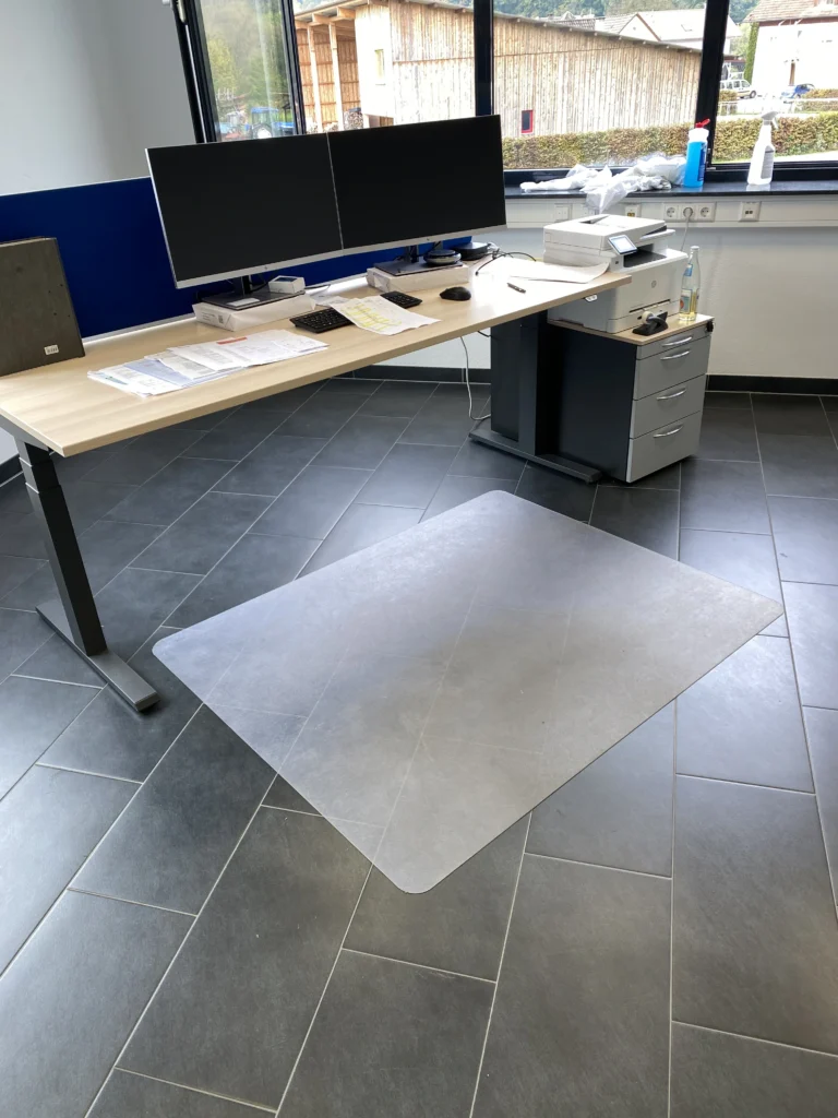 Modernes Büro mit höhenverstellbarem Schreibtisch, zwei Monitoren, einem Drucker und einer Bodenschutzmatte für den Bürostuhl.